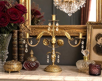 May include: A gold-toned, ornate candelabra with crystal accents is the focal point of this image. The candelabra is surrounded by antique decor, including books, a framed portrait, and a decorative box. A bouquet of red roses adds a pop of color.