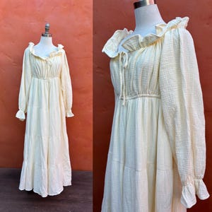 May include: A long-sleeved, floor-length dress in a pale cream color, featuring a textured fabric, a ruffled neckline with a drawstring tie, an elasticized waist, and tiered skirt sections with ruffled cuffs at the end of the long sleeves.