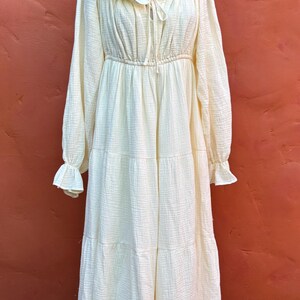 May include: A long-sleeved, floor-length dress in a pale cream color. The garment features a gathered neckline with a tie detail, ruffled edges at the collar and cuffs, and a tiered skirt design. The fabric has a visible, crinkled, textured finish.