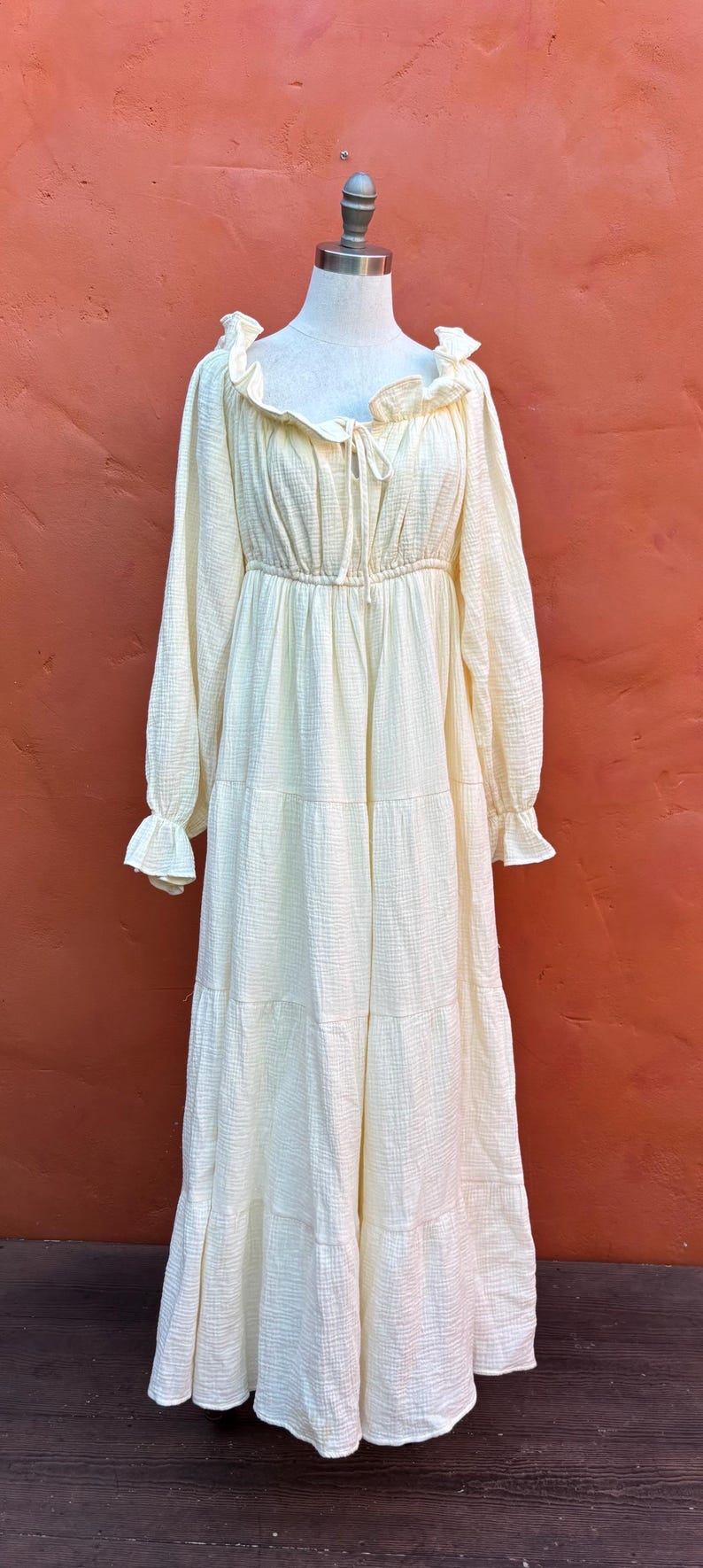May include: A long-sleeved, floor-length dress in a pale cream color. The garment features a gathered neckline with a tie detail, ruffled edges at the collar and cuffs, and a tiered skirt design. The fabric has a visible, crinkled, textured finish.