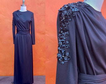 Può includere: Un abito maxi nero con maniche lunghe e un design floreale di paillettes su una spalla. L'abito ha un corpetto aderente e una gonna fluida.