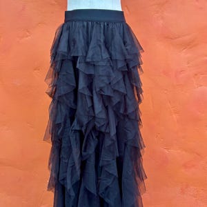 May include: Black tulle skirt with multiple layers of ruffles. The skirt is displayed on a mannequin against an orange background. The skirt has a black waistband.