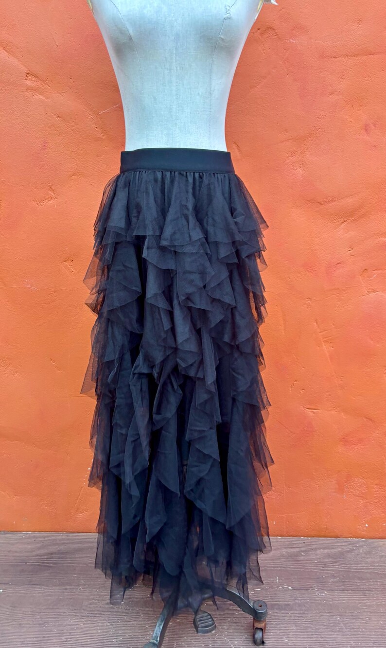 May include: Black tulle skirt with multiple layers of ruffles. The skirt is displayed on a mannequin against an orange background. The skirt has a black waistband.