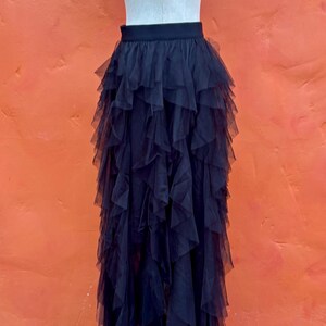 May include: A black tulle skirt with multiple layers of ruffled fabric. The skirt is displayed on a mannequin against an orange background. The skirt's design creates a voluminous, flowing silhouette.