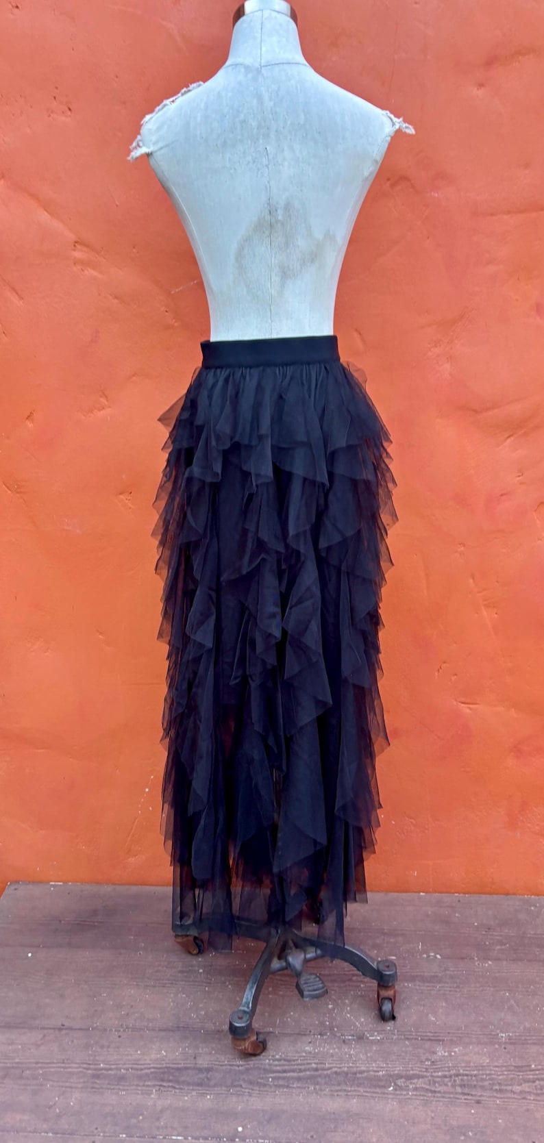 May include: A black tulle skirt with multiple layers of ruffled fabric. The skirt is displayed on a mannequin against an orange background. The skirt's design creates a voluminous, flowing silhouette.