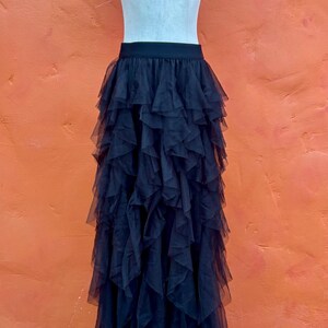 May include: Black tulle skirt with multiple layers of ruffles, displayed on a mannequin. The skirt has a fitted waistband and a full, flowing silhouette. The mannequin is set against an orange wall and a wooden floor.