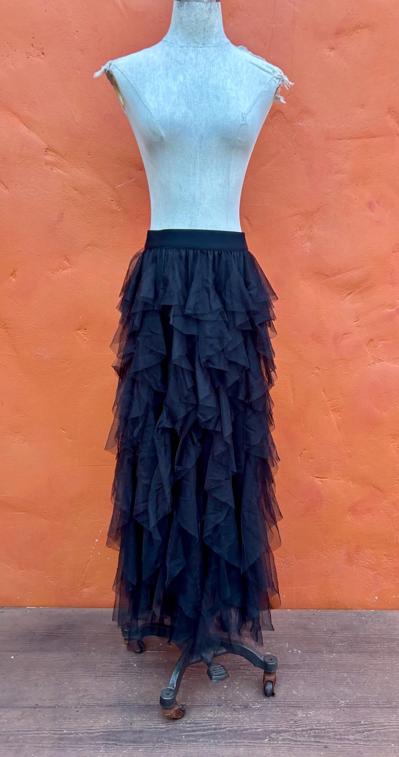 May include: Black tulle skirt with multiple layers of ruffles, displayed on a mannequin. The skirt has a fitted waistband and a full, flowing silhouette. The mannequin is set against an orange wall and a wooden floor.