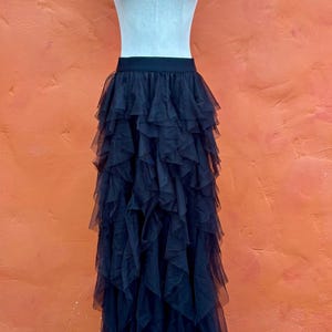 May include: A black tulle skirt with multiple layers of ruffled fabric, displayed on a dress form. The skirt has a wide, elastic waistband. The background is a solid orange wall.