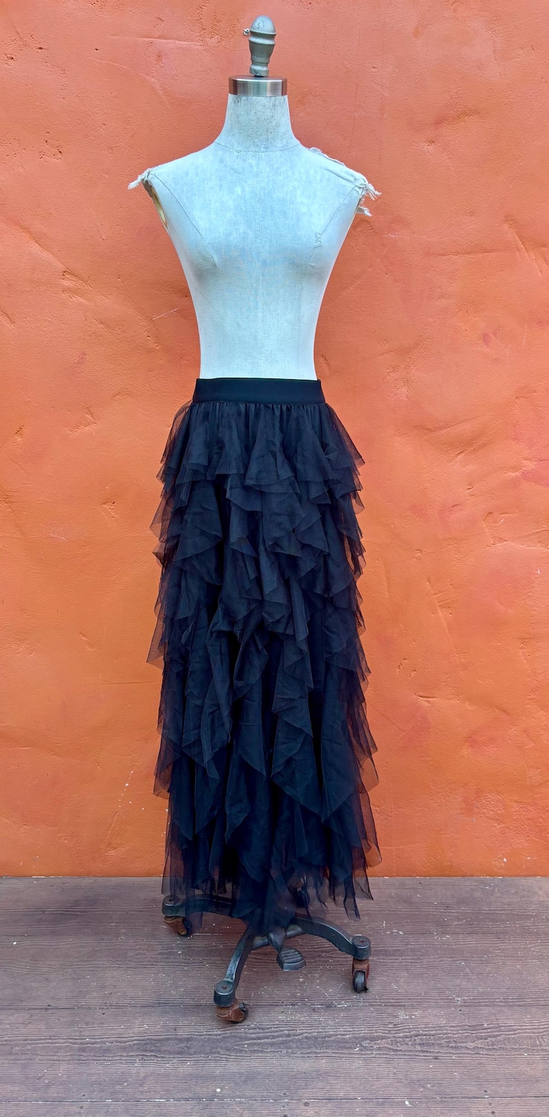 May include: A black tulle skirt with multiple layers of ruffled fabric, displayed on a dress form. The skirt has a wide, elastic waistband. The background is a solid orange wall.