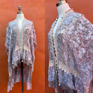 以下が含まれることがあります： ラベンダー、グレー、クリーム色の花柄のシアーな着物。マネキンに展示されています。着物には、縁に繊細なレースのトリムとフリルの裾が付いています。この衣服は軽く、流れるような、ボヘミアンな美学を持っています。