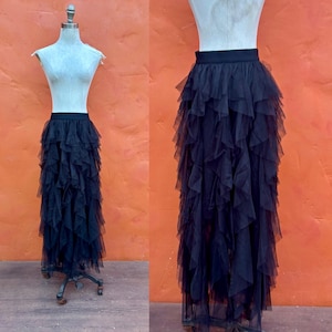 May include: A black tulle skirt with multiple layers of ruffles, displayed on a mannequin. The skirt has a fitted waistband and a floor-length hemline. The mannequin is set against an orange background.