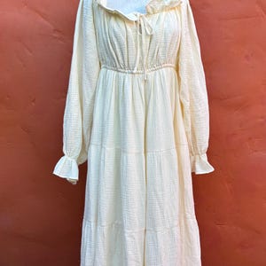 May include: A long-sleeved, floor-length dress in a pale cream color. The garment features a gathered neckline with a tie detail, ruffled edges at the collar and cuffs, a cinched elastic waist, and a tiered skirt made of a textured fabric.