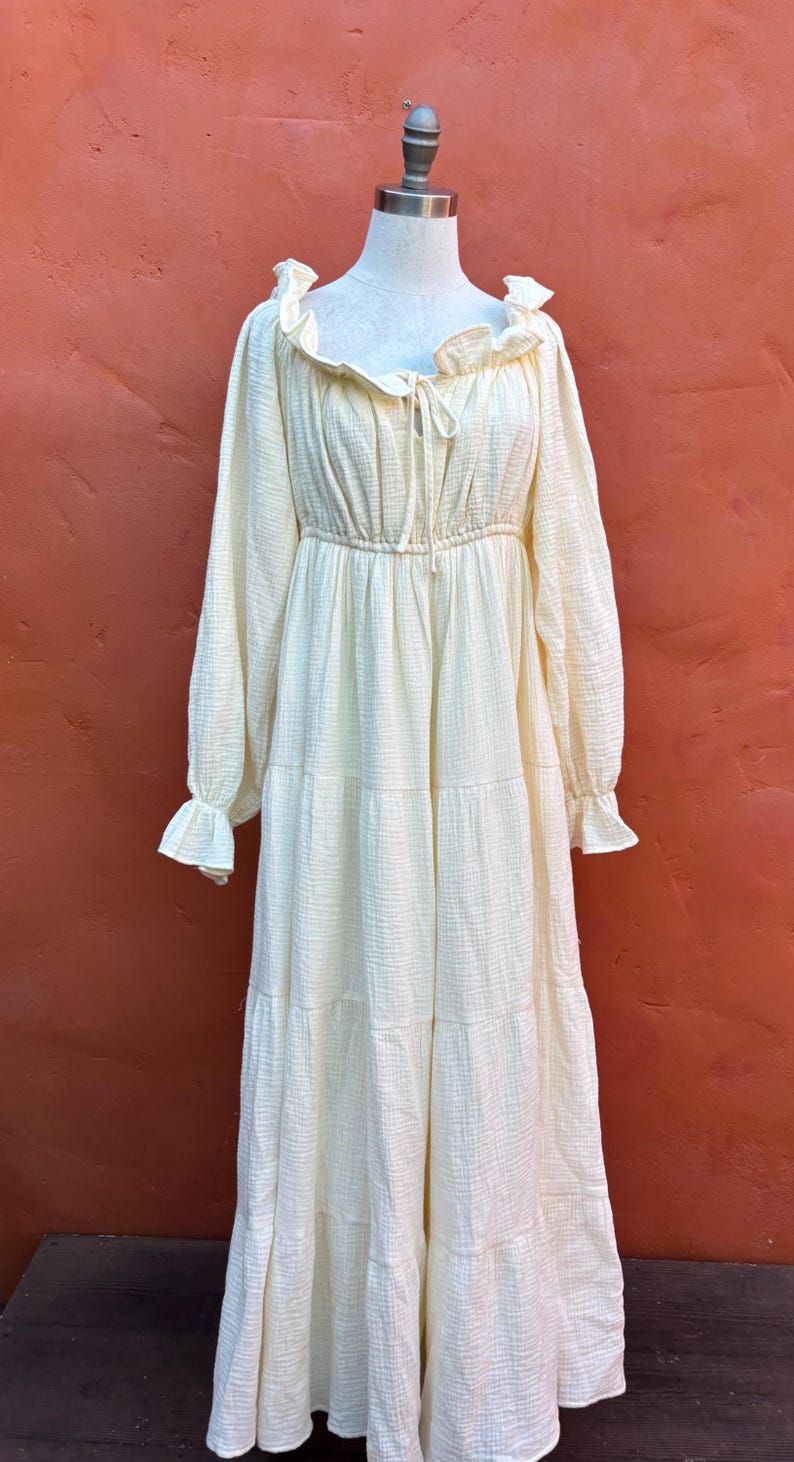 May include: A long-sleeved, floor-length dress in a pale cream color. The garment features a gathered neckline with a tie detail, ruffled edges at the collar and cuffs, a cinched elastic waist, and a tiered skirt made of a textured fabric.
