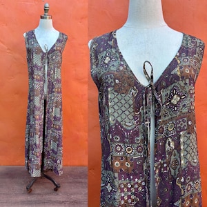 May include: A sleeveless, floor-length duster vest featuring a deep V-neckline with a thin tie closure at the center. The garment is decorated with a multi-colored patchwork pattern in shades of deep purple, olive green, brown, and cream.