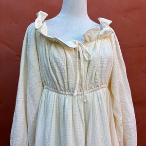 May include: A pale yellow long-sleeved dress with a textured, crinkled fabric finish. The garment features a ruffled neckline, a drawstring tie at the center chest, and an elasticized waistline that creates a gathered, flowing silhouette.