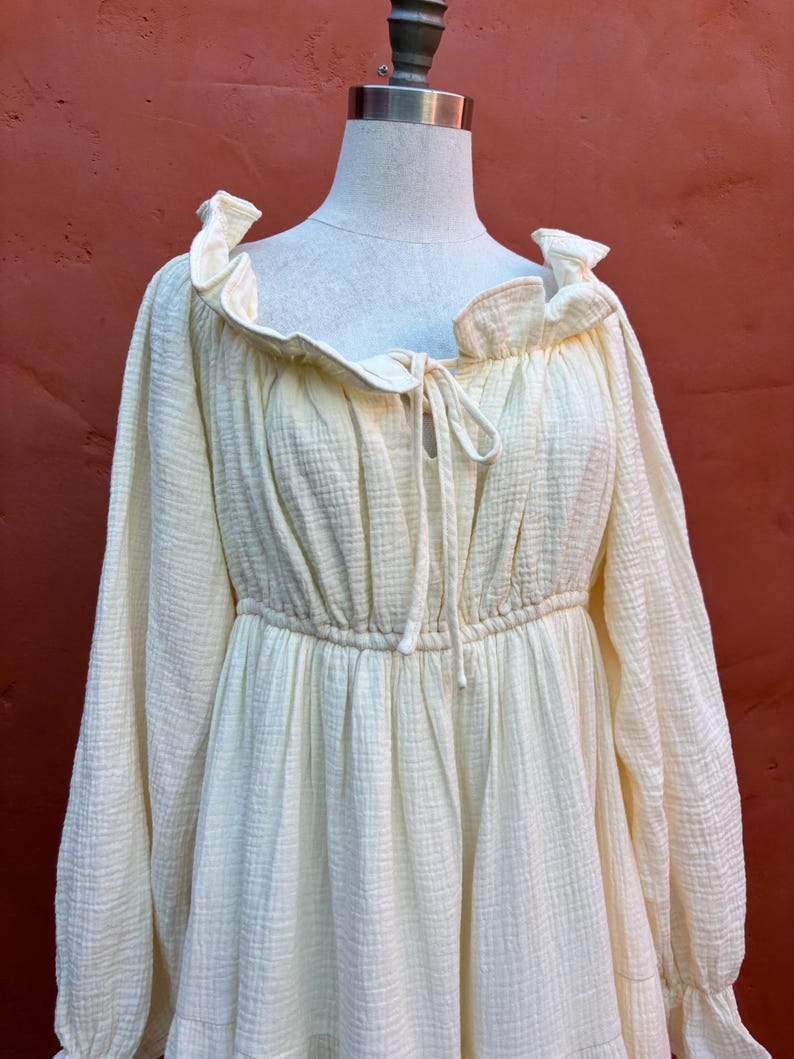 May include: A pale yellow long-sleeved dress with a textured, crinkled fabric finish. The garment features a ruffled neckline, a drawstring tie at the center chest, and an elasticized waistline that creates a gathered, flowing silhouette.