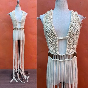 May include: Cream-colored macrame top and skirt set displayed on a mannequin. The top features a deep V-neck and a cut-out design. The skirt has long fringe detailing, creating a bohemian aesthetic. The set is made of woven cotton.