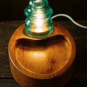 May include: A unique table lamp featuring a teal glass insulator atop a wooden base. The base is a rounded, wooden bowl shape with a carved-out section. The lamp is lit, emitting a warm glow.