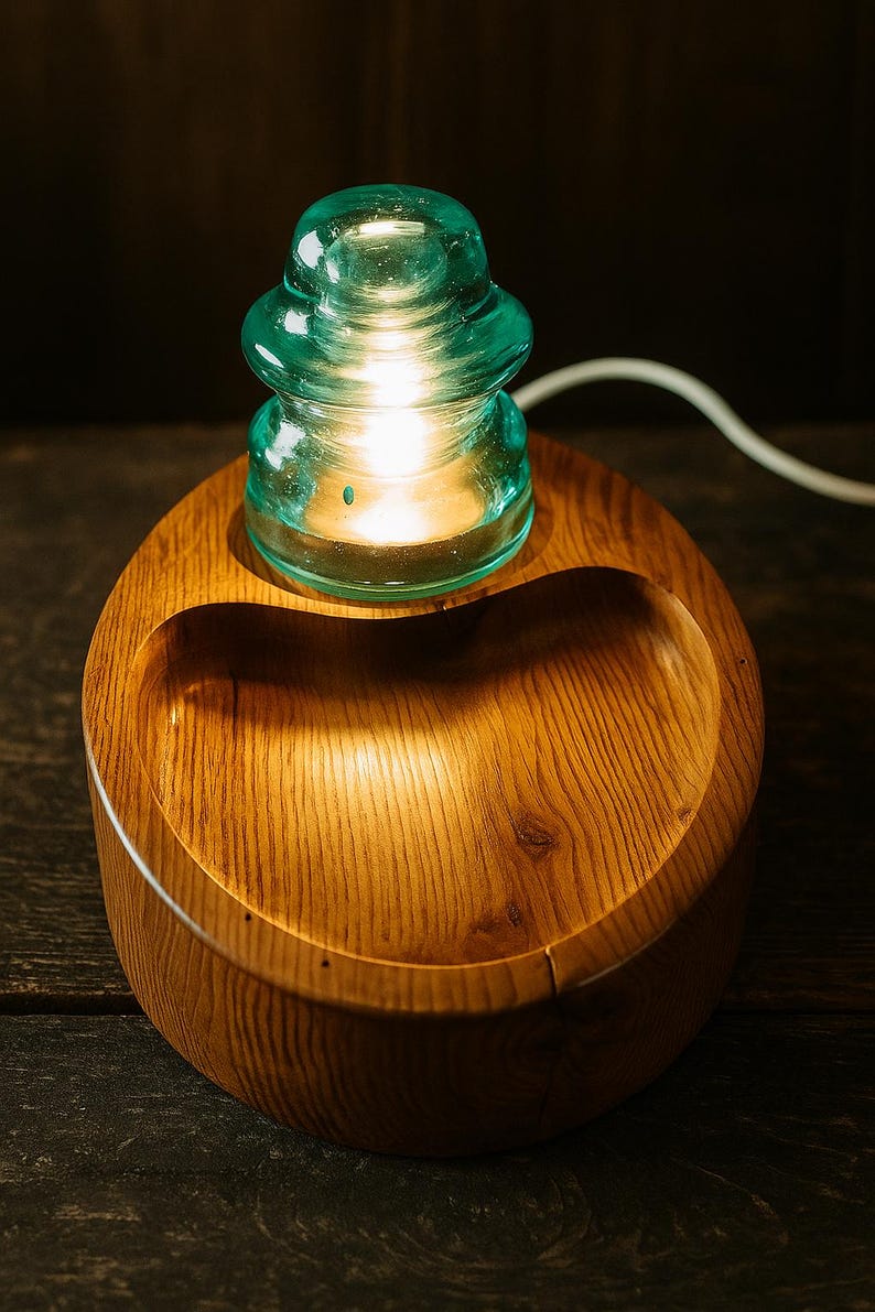 May include: A unique table lamp featuring a teal glass insulator atop a wooden base. The base is a rounded, wooden bowl shape with a carved-out section. The lamp is lit, emitting a warm glow.