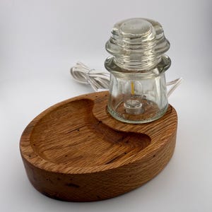 May include: A unique lamp featuring a clear glass insulator with a small light bulb, set on a handcrafted, oval-shaped wooden base. The base has a carved-out section and a white electrical cord.