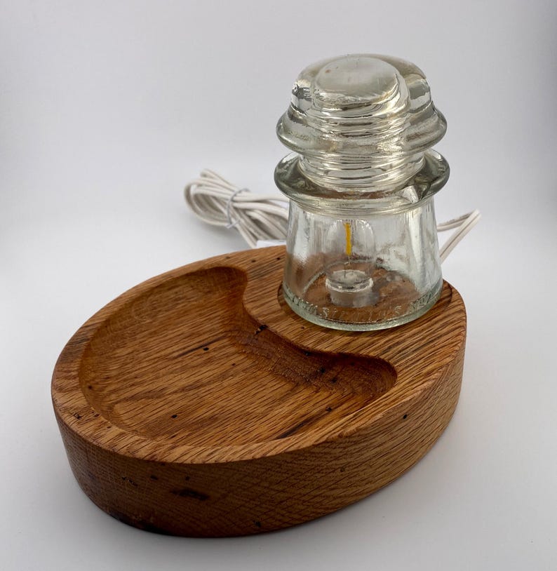 May include: A unique lamp featuring a clear glass insulator with a small light bulb, set on a handcrafted, oval-shaped wooden base. The base has a carved-out section and a white electrical cord.