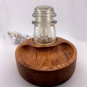 May include: A unique table lamp featuring a clear glass insulator with a small light bulb, set on a round, wooden base. The base is made of a dark, textured wood. A white electrical cord with a plug extends from the lamp.