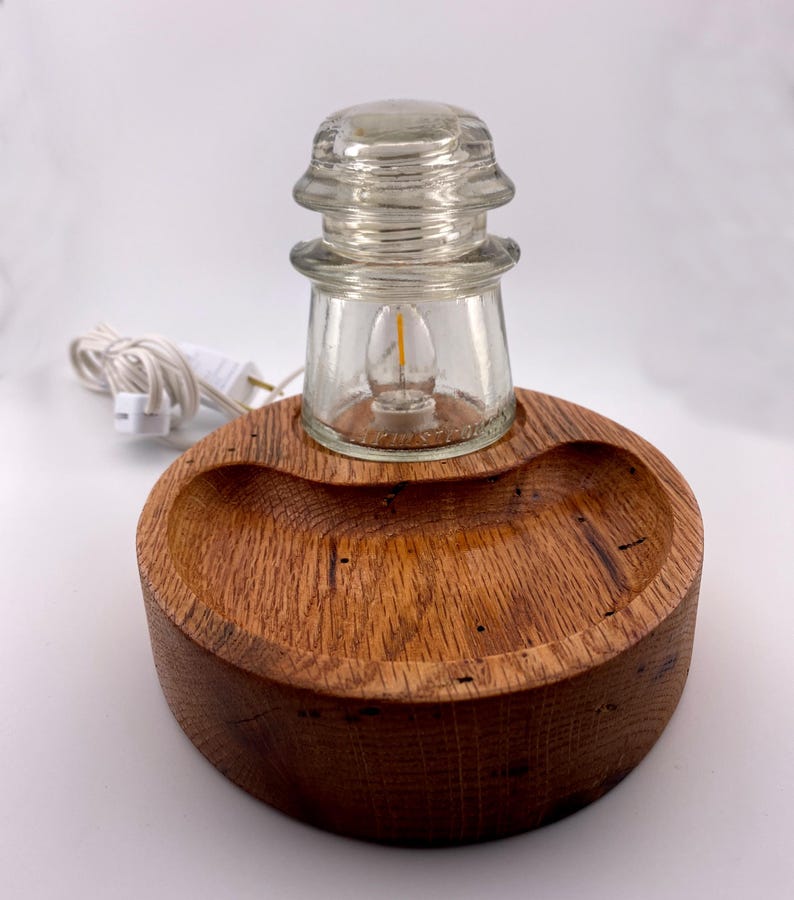 May include: A unique table lamp featuring a clear glass insulator with a small light bulb, set on a round, wooden base. The base is made of a dark, textured wood. A white electrical cord with a plug extends from the lamp.