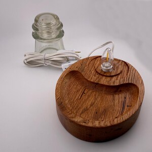 May include: A handcrafted wooden lamp base with a small light bulb and a clear glass insulator. The lamp base is round with a curved groove and a natural wood grain finish. A white power cord is attached.