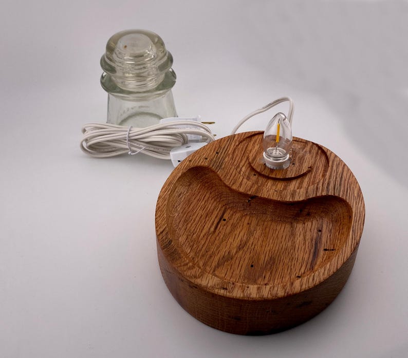 May include: A handcrafted wooden lamp base with a small light bulb and a clear glass insulator. The lamp base is round with a curved groove and a natural wood grain finish. A white power cord is attached.