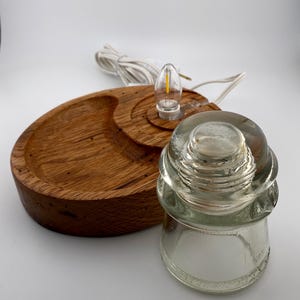May include: A handcrafted wooden lamp base with a clear glass insulator and a small LED light bulb. The base is made of light brown wood and has a circular design. The lamp includes a white electrical cord.