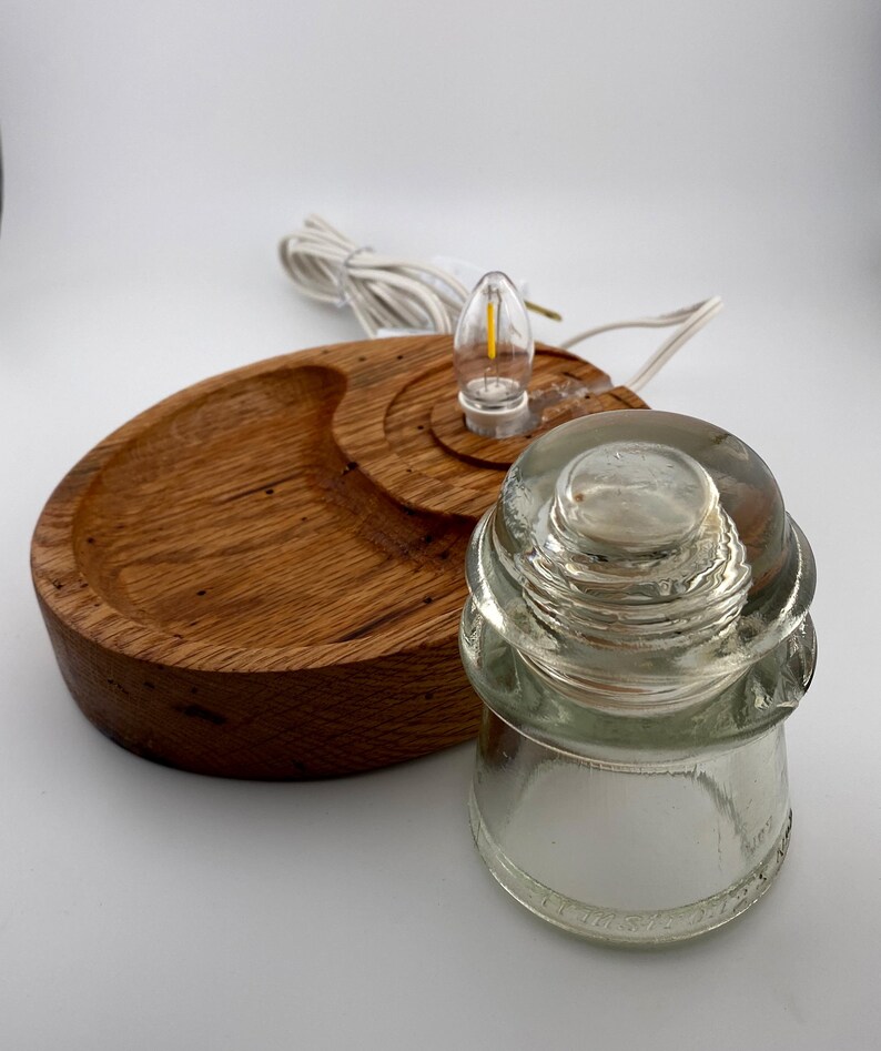May include: A handcrafted wooden lamp base with a clear glass insulator and a small LED light bulb. The base is made of light brown wood and has a circular design. The lamp includes a white electrical cord.