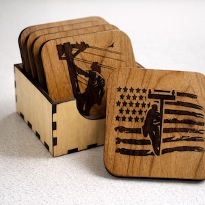 May include: Set of wooden coasters with laser-etched designs. One coaster features an American flag design with a silhouette of a person. The other coasters depict a person working on a power line. The coasters are stored in a wooden box.