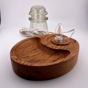 May include: A handcrafted wooden lamp with a warm, natural finish. The lamp features an oval base with a carved design, a small light bulb, and a clear glass insulator. The lamp is powered by a white cord.
