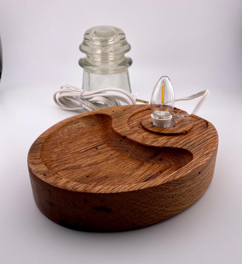 May include: A handcrafted wooden lamp with a warm, natural finish. The lamp features an oval base with a carved design, a small light bulb, and a clear glass insulator. The lamp is powered by a white cord.
