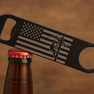 May include: Black bottle opener with a handle and a circular opening. The opener features an American flag design and a silhouette of a lineman climbing a utility pole. The opener is resting on a brown glass bottle with a red cap.