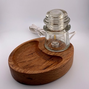 May include: A unique lamp featuring a clear glass insulator with a small light bulb, set on a wooden base. The base is oval-shaped with a carved-out section. A white electrical cord extends from the lamp.