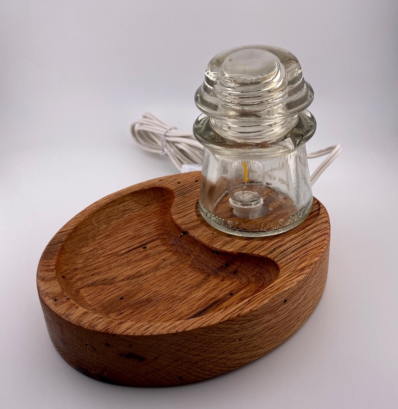 May include: A unique lamp featuring a clear glass insulator with a small light bulb, set on a wooden base. The base is oval-shaped with a carved-out section. A white electrical cord extends from the lamp.