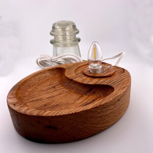 May include: A handcrafted wooden lamp with a warm, natural finish. The lamp features a unique, oval-shaped base with a carved design and a small light bulb. A clear glass insulator and white cord are also visible.