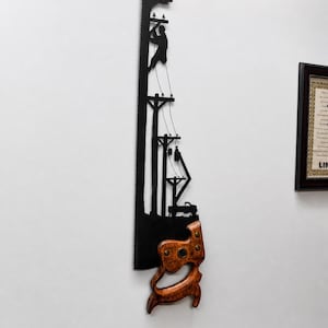 May include: A black metal hand saw wall art piece featuring a cutout silhouette of a utility worker climbing a power pole, with additional utility poles, power lines, and a service truck depicted along the blade, finished with a wooden handle.