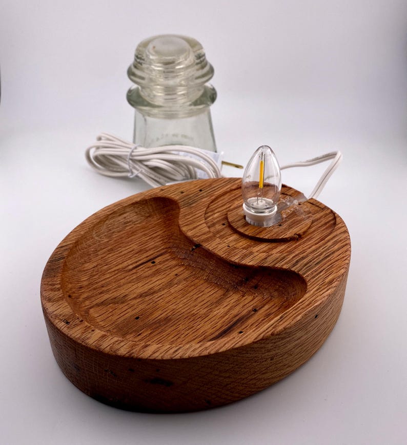 May include: A handcrafted wooden lamp base with a clear glass insulator and a small LED light. The base is oval-shaped with a carved dish and a circular area for the light. The wood has a warm, natural finish.