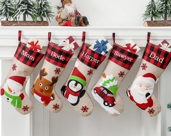 May include: Five personalized Christmas stockings with red and black plaid trim. The stockings feature different characters, including a snowman, reindeer, penguin, car, and Santa Claus. The stockings are labeled with the names "Mom", "Bode", "Harper", "Sawyer", and "Dad".