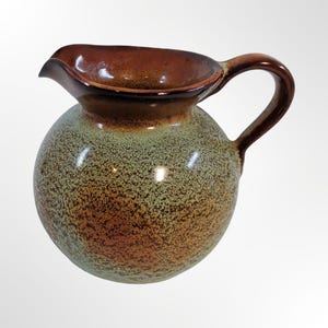 May include: A round ceramic pitcher featuring a speckled sage green and brown finish. The vessel has a wide, bulbous body, a curved handle on the side, and a prominent pouring spout at the top rim, which is glazed in a solid, glossy dark brown.