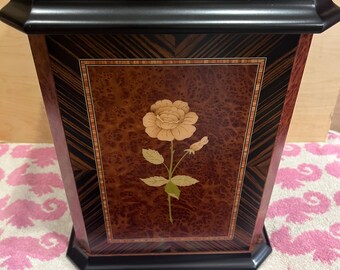 May include: A decorative wooden box with a floral inlay. The box features a rectangular shape with a dark wood frame and a lighter wood top. The front panel showcases a detailed rose design in cream and green.