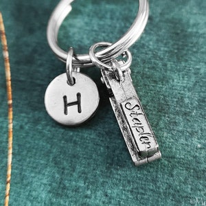 May include: Silver keychain with a letter H charm and a charm shaped like a stapler with the word "Stapler" engraved on it.