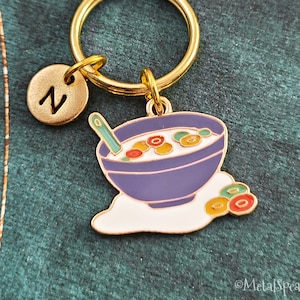 May include: Gold keychain with a purple bowl of cereal and a spoon. The bowl is filled with white cereal and colorful cereal pieces. There are three colorful cereal pieces on the white surface below the bowl. The keychain has a gold letter "N" charm.