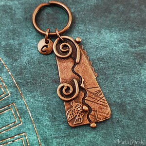 Peut inclure: Un porte-clés en cuivre avec un charme décoratif représentant un motif en spirale, un oiseau et un petit charme circulaire avec la lettre "C".