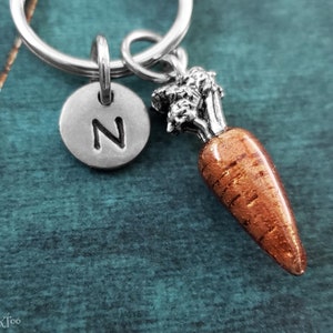 May include: A silver keychain with a carrot charm and a silver charm with the letter N. The carrot charm is a detailed representation of a carrot with a green top and an orange body.