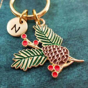 May include: Gold-tone keyring with a green pine branch, brown pine cone, and red berries. The keyring also features a gold-tone initial charm with the letter "N".
