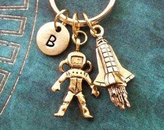 May include: Gold-toned keychain with a letter B charm, a small astronaut charm, and a space shuttle charm. The charms are attached to a gold-toned key ring.