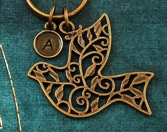 May include: A bronze-colored metal keychain featuring a dove design with intricate cut-out details. The dove has a small circular charm with the letter "A" and is attached to a round key ring. The background is a teal-colored fabric.